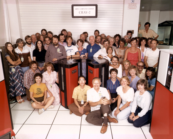 About 50 people sit and stand around a barrel-shaped red and black  Cray-2 supercomputer.