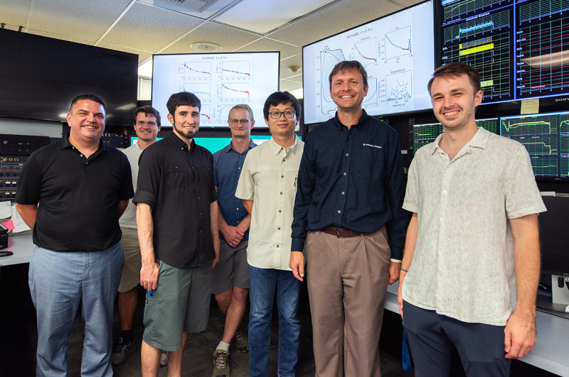 Team of smiling engineers stand in front of a bank of monitors showing various line graphs and monitoring dashboards.