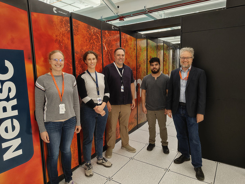 Two women and three men stand smiling before Perlmutter's colorful supercomputer cabinets.