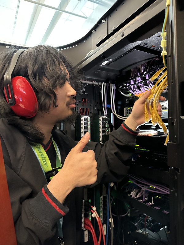 A man with long hair and hearing protection inspects cables inside of a supercomputer's cabinet.
